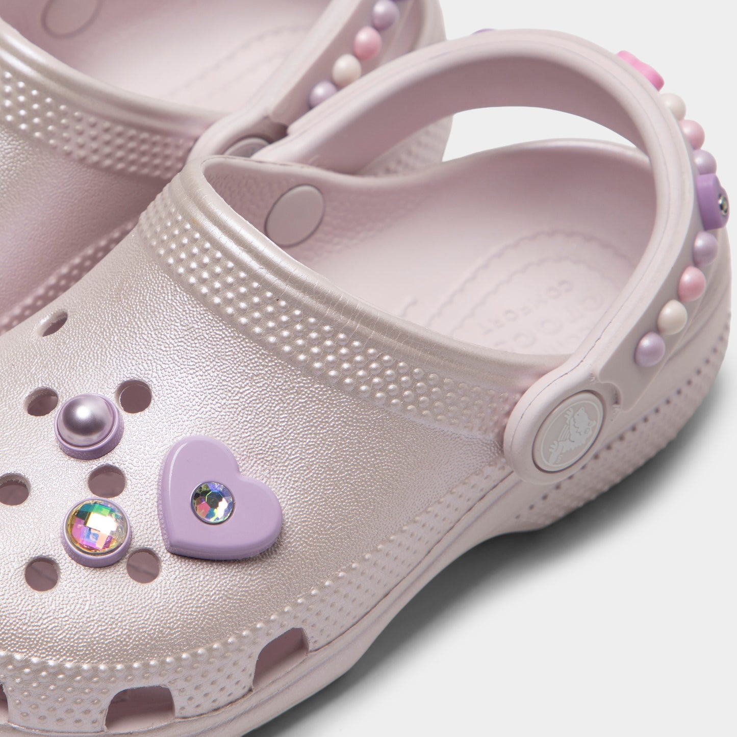 Crocs Kids' Classic Pearls and Heart Shine Clog. Close-up of grape ice infant clog showing pivoting heel strap, pearl and heart charms and Crocs logo button.