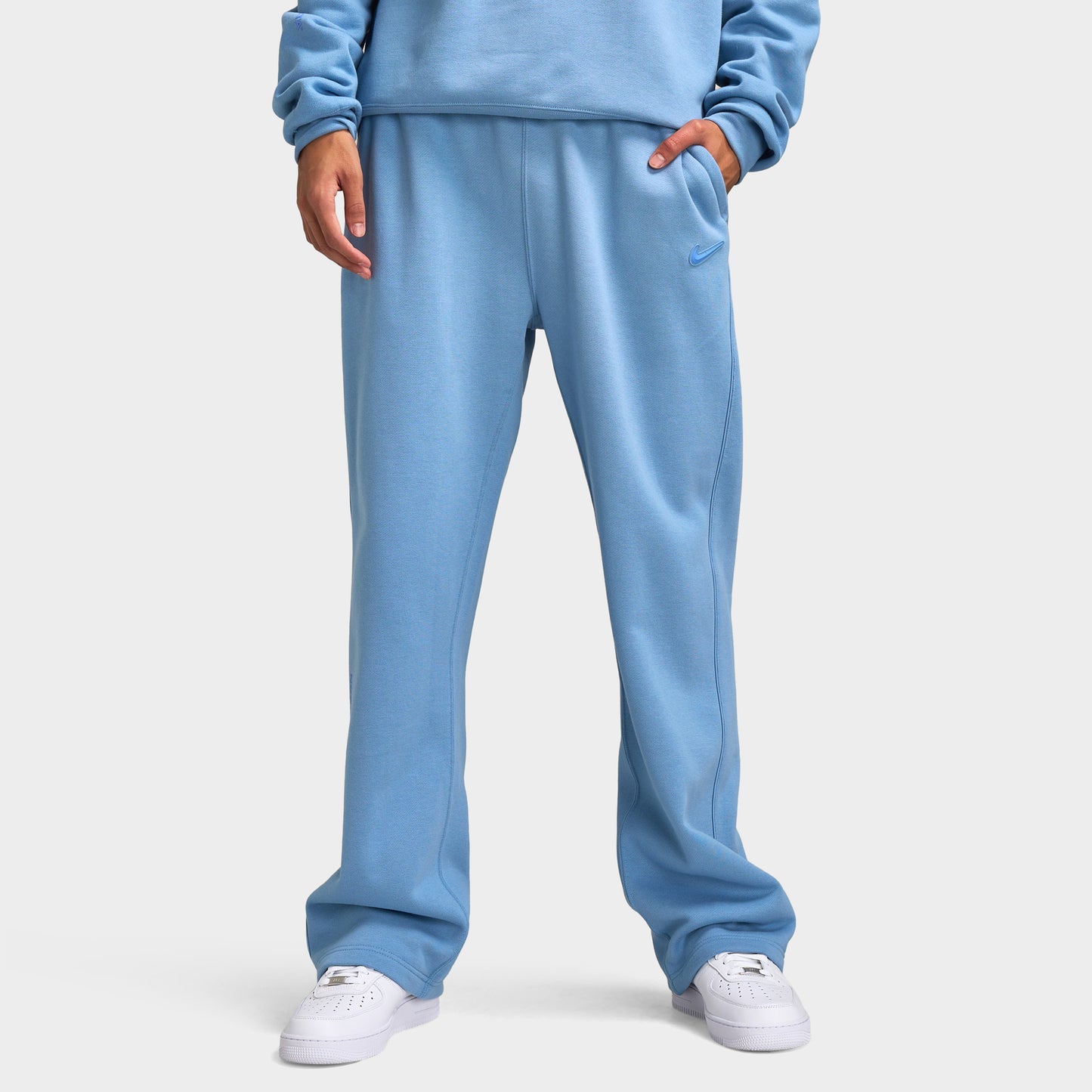 Nike NOCTA Fleece Open Hem Pants. Front view of model wearing work blue sweatpants with wide legs, elastic waistband and side pockets.