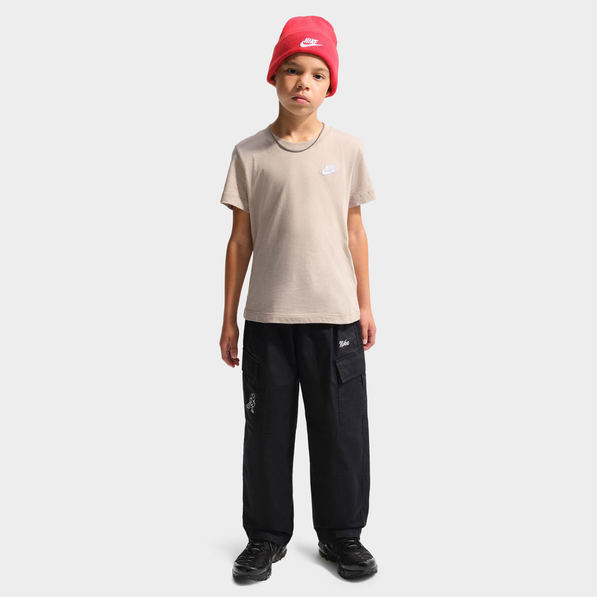 Nike Juniors' Futura T-Shirt. Full-length front view of a youth model wearing a red beanie and the moon particle beige shortsleeve t-shirt paired with black trousers and black sneakers.