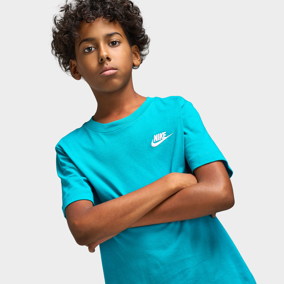 Nike Juniors' Futura T-Shirt. Three-quarter upper body model view displaying energy green turquoise shortsleeve t-shirt with arms crossed, ribbed crew collar, and white embroidered Nike Futura chest logo.