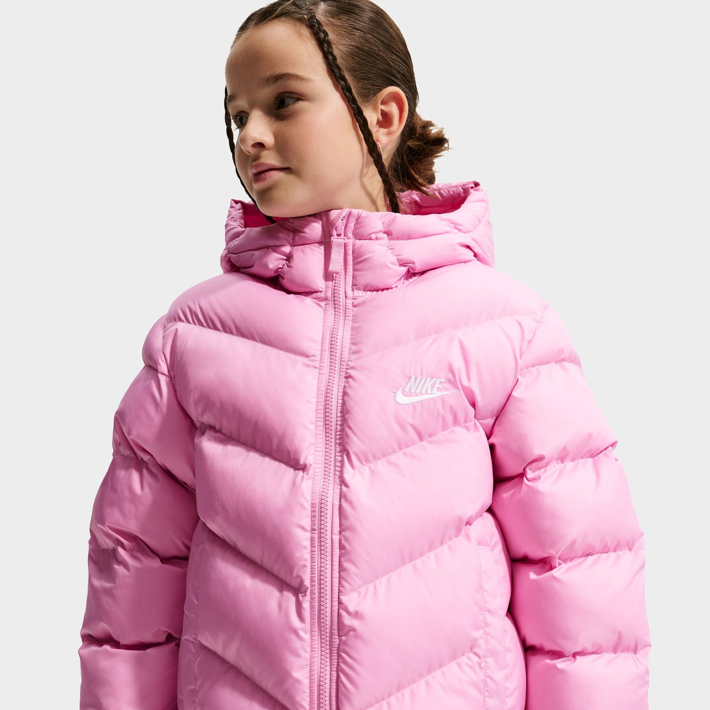 Nike Junior Girls' Therma-FIT Loose-Fit Puffer Jacket. Three-quarter view of model wearing pink rise puffer jacket with hood, zip closure and quilted sleeves.