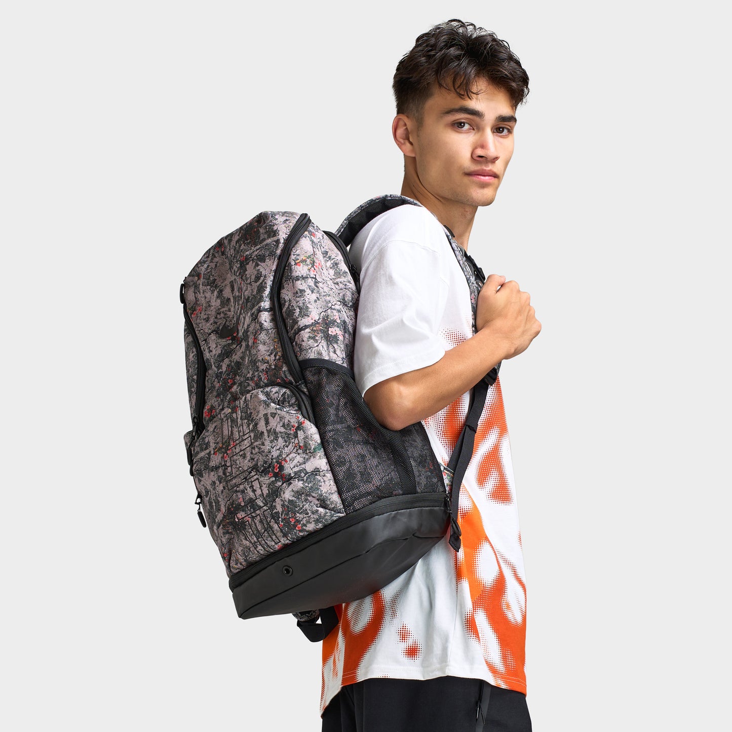 Nike Varsity Elite Backpack. Three-quarter view of model wearing cream and black backpack with all-over graphic print, padded shoulder straps and structured base.