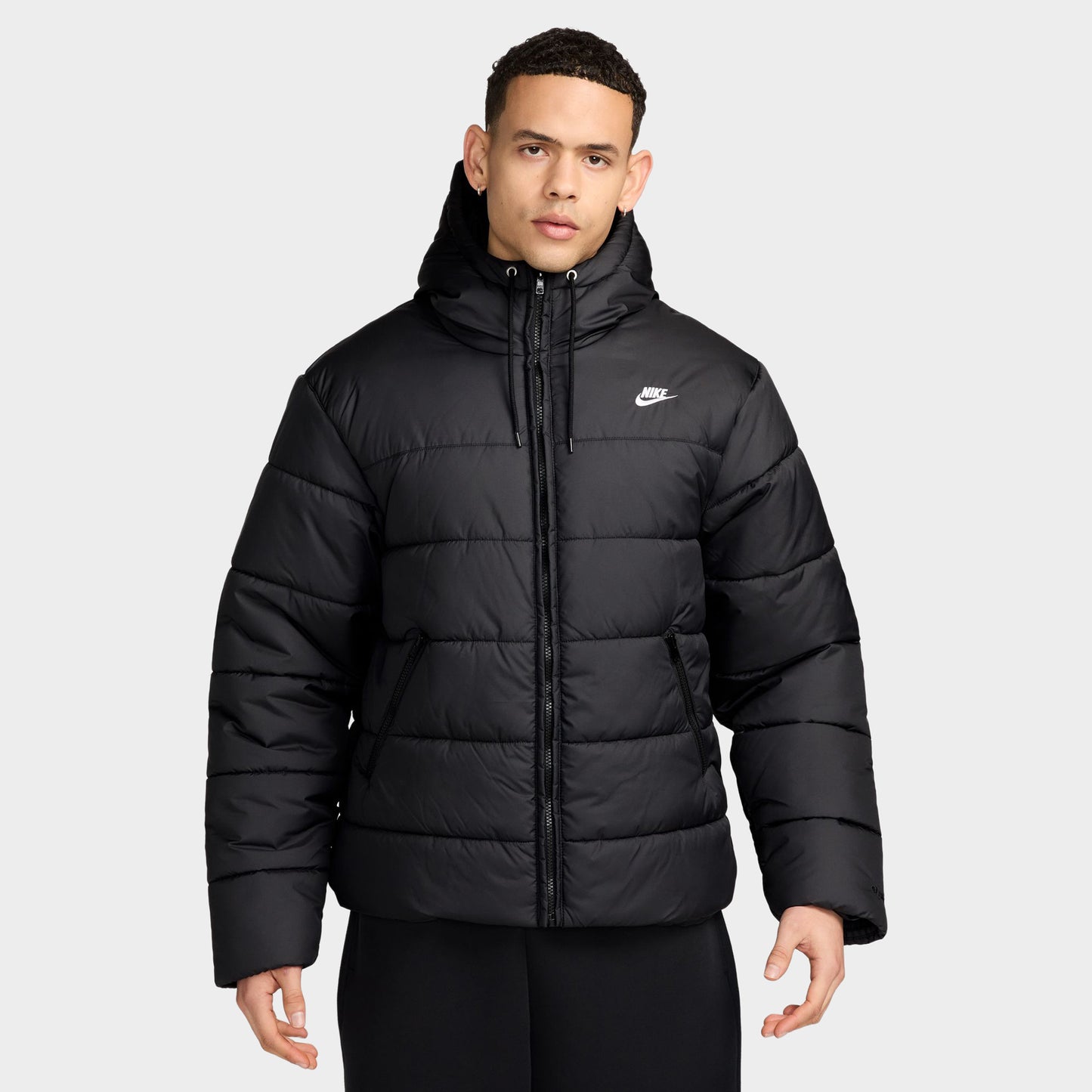 Nike Access Filled Puffer. Front view of model wearing black puffer jacket with hood, zip closure, quilted panels and white Nike Swoosh.