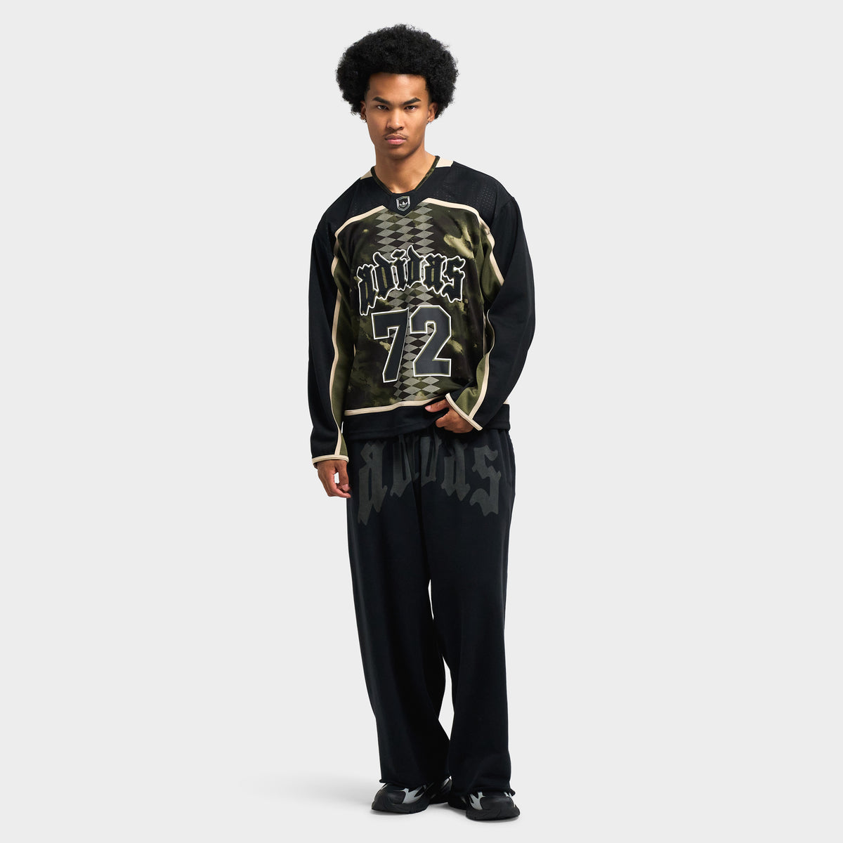 adidas Hockey Jersey. Full-length rear view of model wearing olive strata camo hockey jersey paired with loose black pants and sneakers.