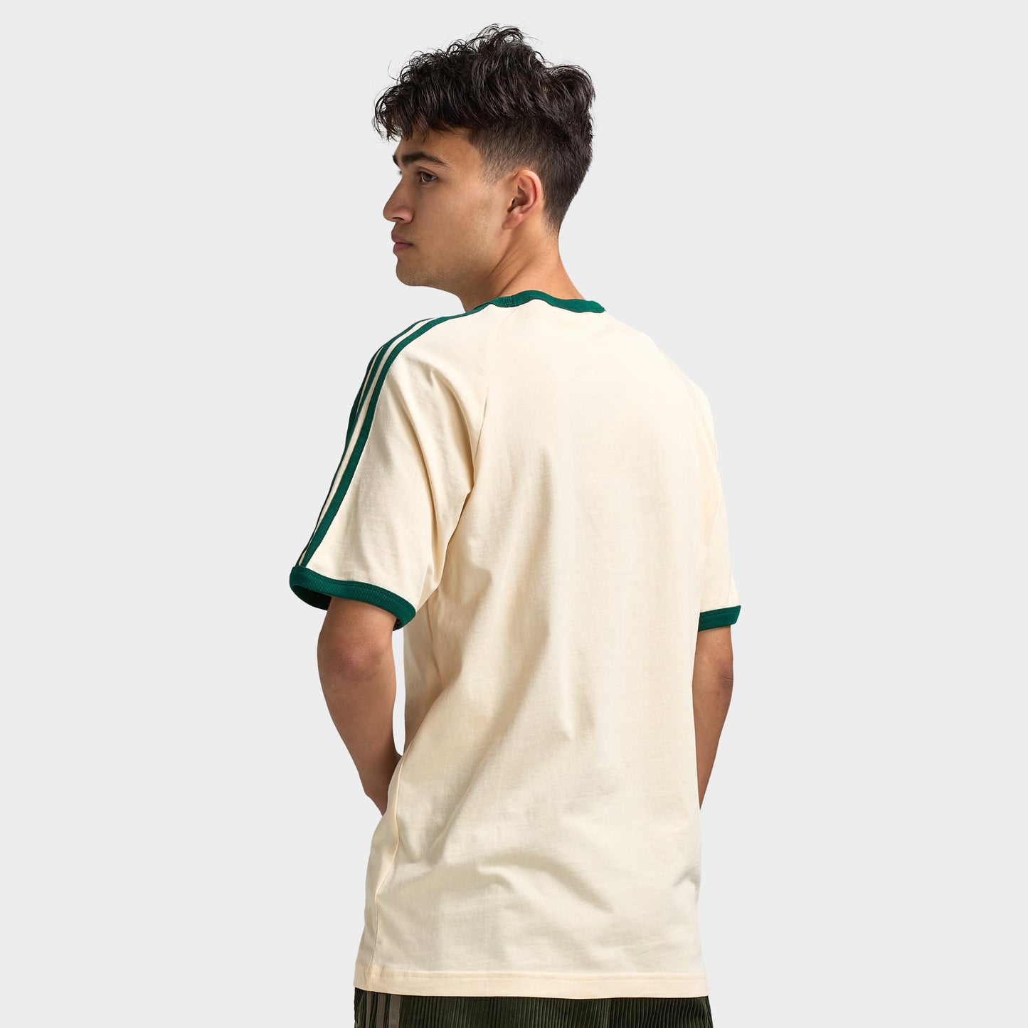 adidas 3 Stripes T-Shirt. Back three-quarter view of model wearing vanilla and collegiate green T-shirt with green sleeve stripes and relaxed fit.