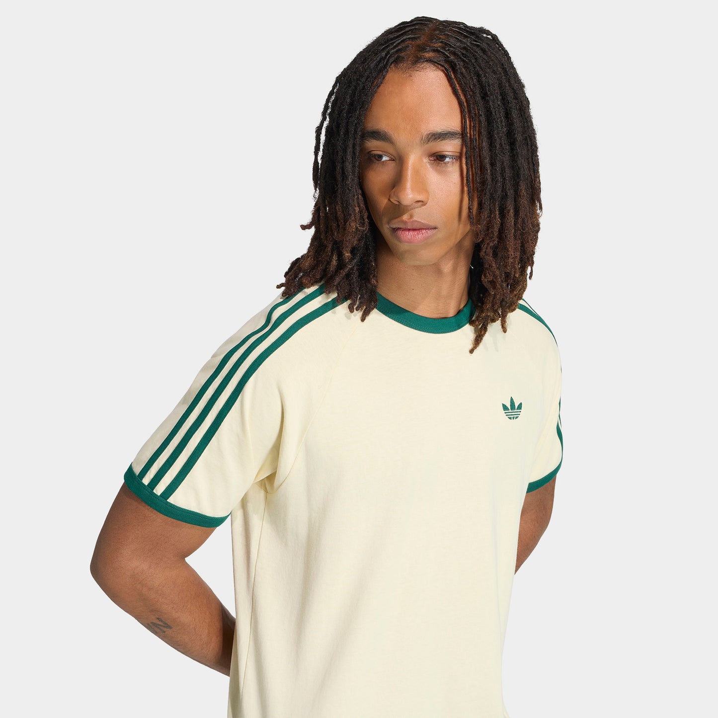 adidas 3 Stripes T-Shirt. Three-quarter view of model wearing vanilla and collegiate green T-shirt with striped sleeves and green Trefoil logo.