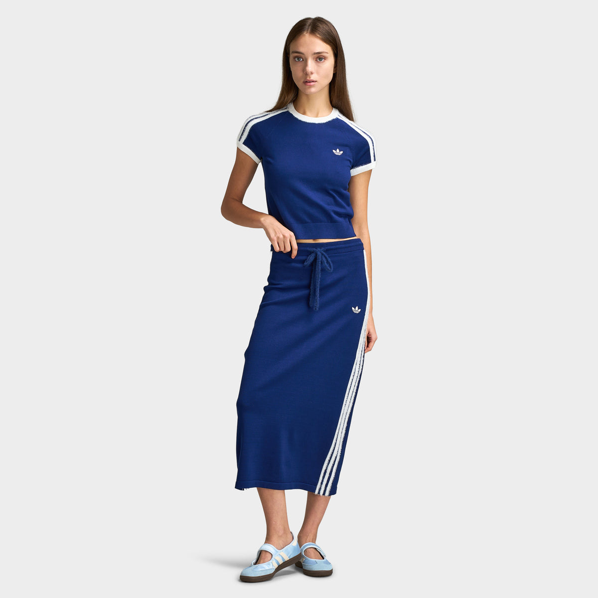 adidas 3 Stripe Knitted Skirt. Full-length front model view styled with a dark blue knitted shortsleeve top with white 3-Stripes on the shoulders, matching dark blue 3-Stripe knitted midi skirt, and light blue Mary Jane shoes.