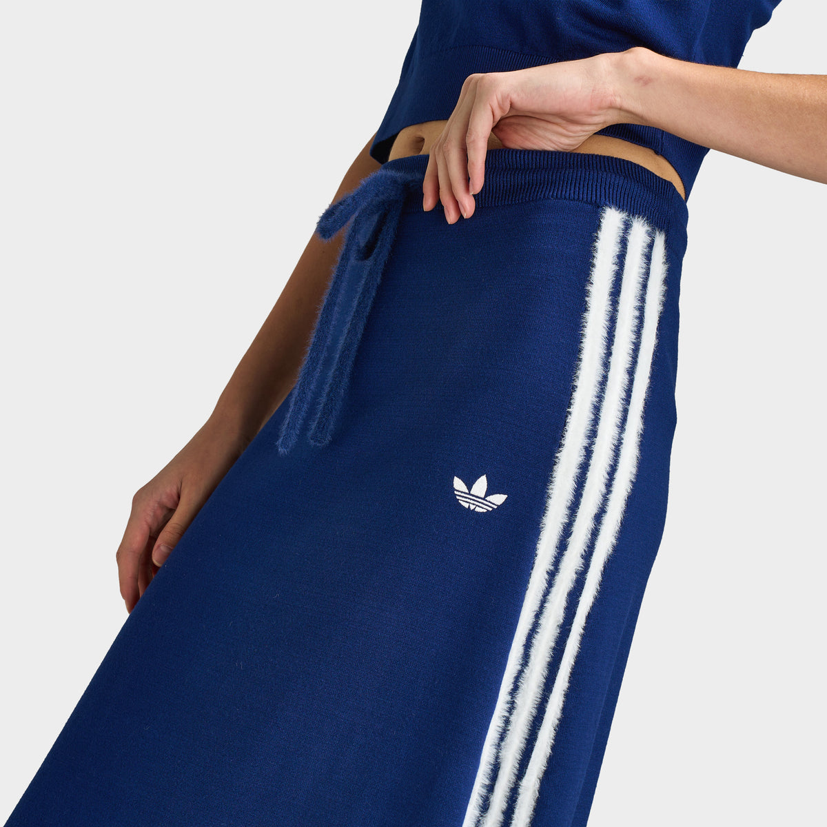 adidas 3 Stripe Knitted Skirt. Close-up view of upper hip highlighting dark blue fine-knitted fabric, elasticated waistband with tonal drawcord, small white embroidered adidas Trefoil logo, and contrast white 3-Stripes side piping.