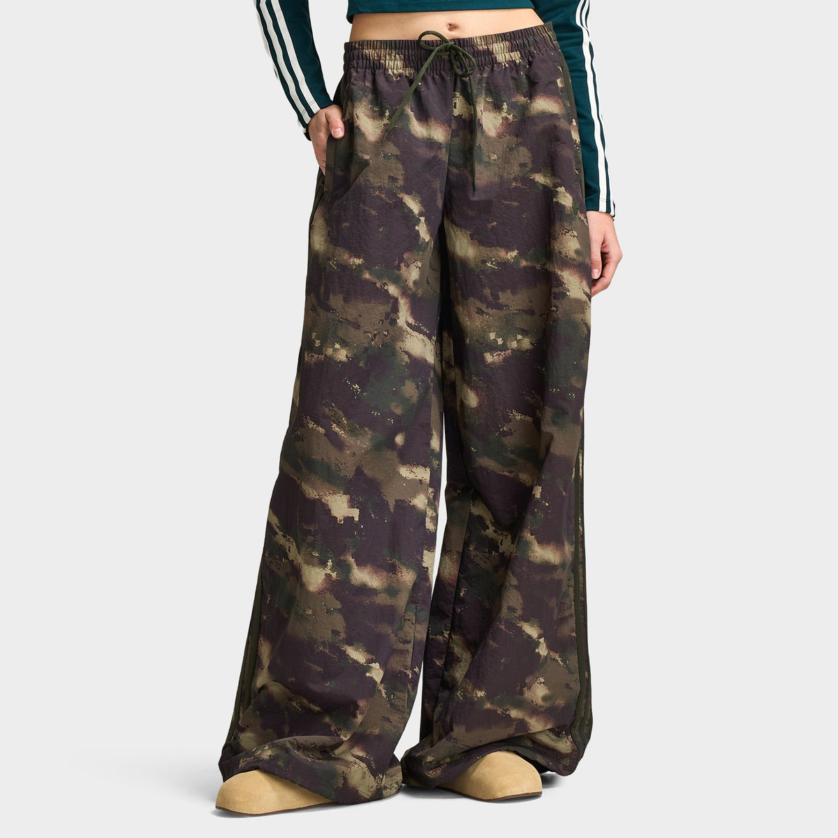 adidas Women's Camouflage Adilenium Track Pant. Lower body front model view of night cargo, aurora coffee, and olive strata camouflage print wide-leg track pants featuring an elasticated waistband with adjustable drawstring, side pockets, tonal 3-Stripes along the side seams, wide-leg loose cut, and floor-length open hem.