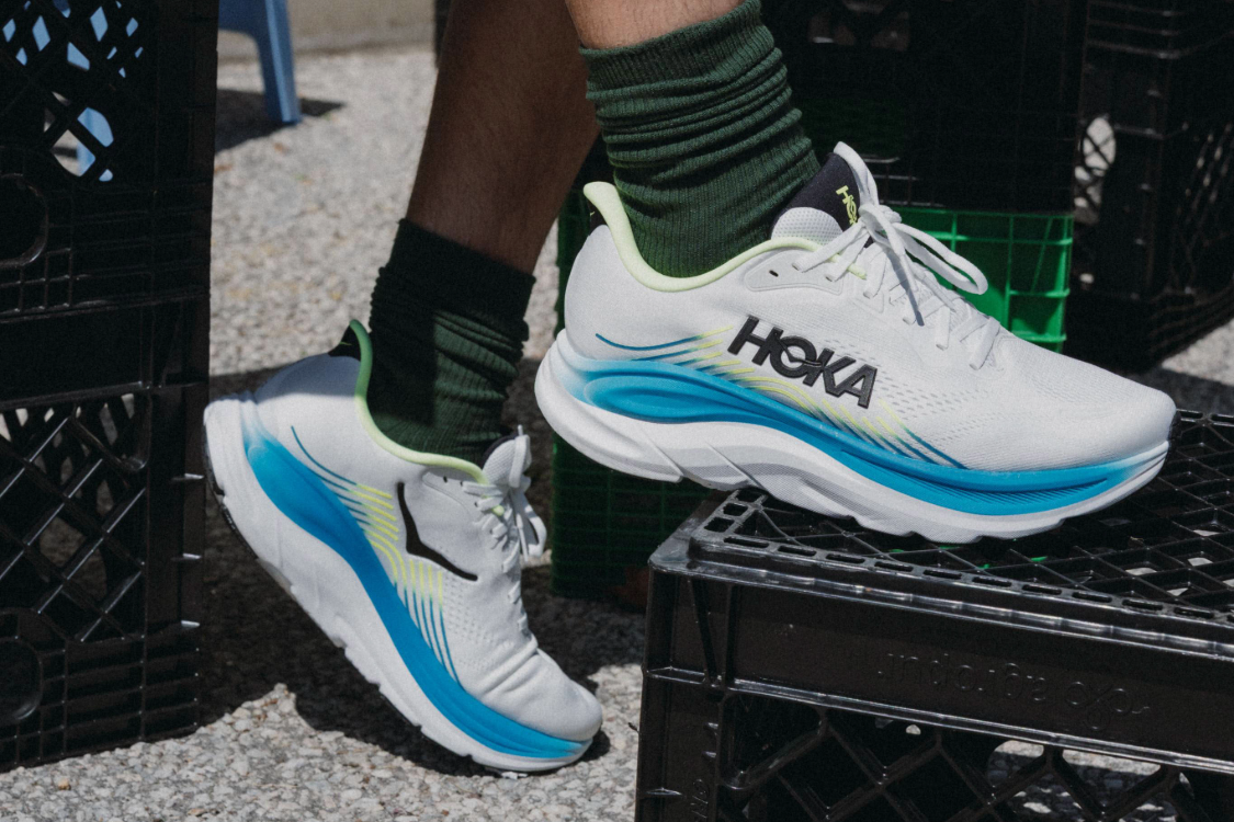 A close-up view of a person wearing white and blue Hoka running shoes with green socks while resting one foot on a black crate.