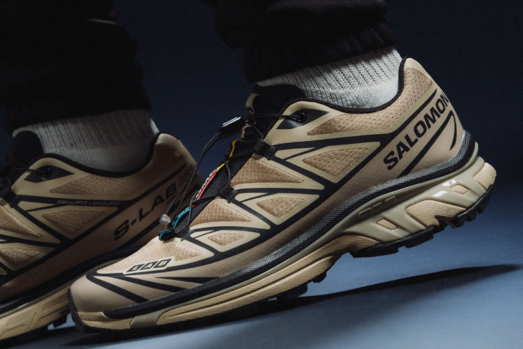 Close-up of tan and black Salomon trail running shoes worn with black pants on a dark background.