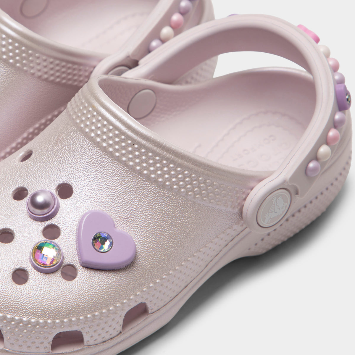 Crocs Kids' Classic Pearls and Heart Shine Clog. Close-up of grape ice infant clog showing pivoting heel strap, pearl and heart charms and Crocs logo button.