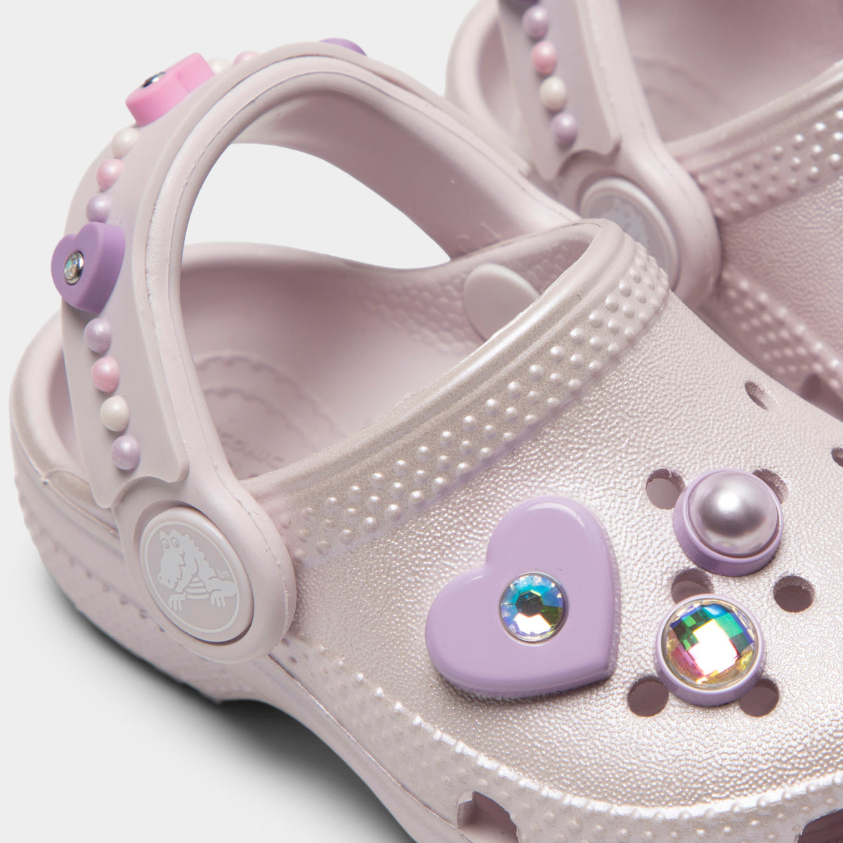 Crocs Infants' Classic Pearls and Heart Shine Clog. Close-up of grape ice infant clog showing pivoting heel strap, pearl and heart charms and Crocs logo button.