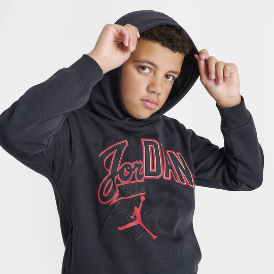 Jordan Juniors' Varsity Split Pullover Hoodie. Three-quarter view of model wearing off noir and gym red hoodie with Jordan chest graphic and sleeve patches.