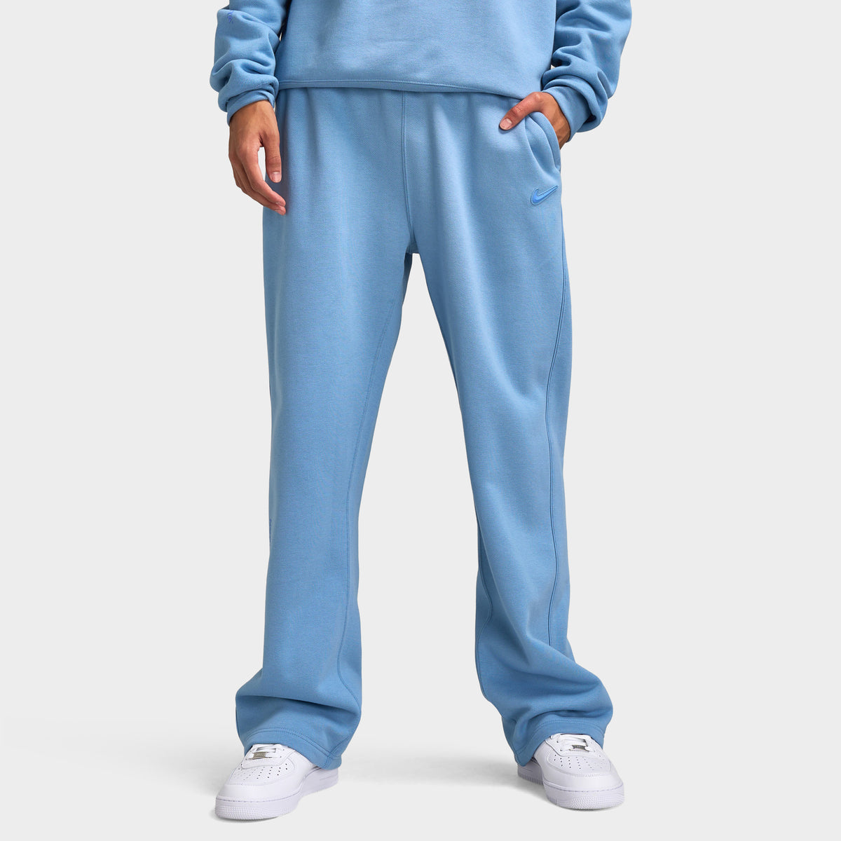 Nike NOCTA Fleece Open Hem Pants. Front view of model wearing work blue sweatpants with wide legs, elastic waistband and side pockets.