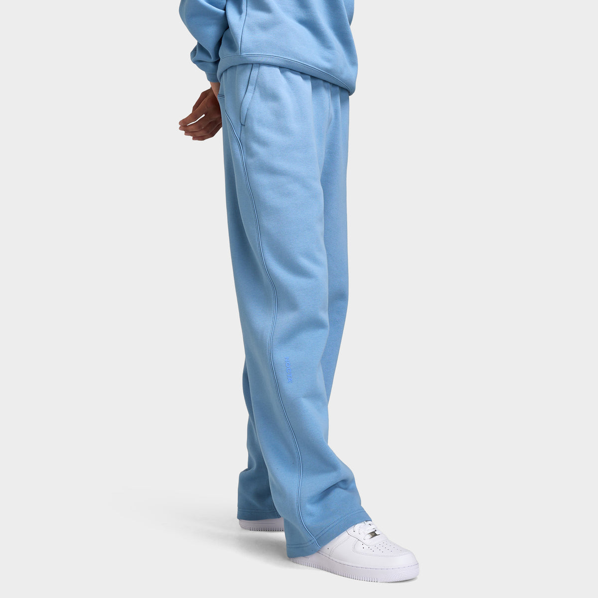 Nike NOCTA Fleece Open Hem Pants. Three-quarter view of model wearing work blue sweatpants with wide legs, side pockets and open hems.