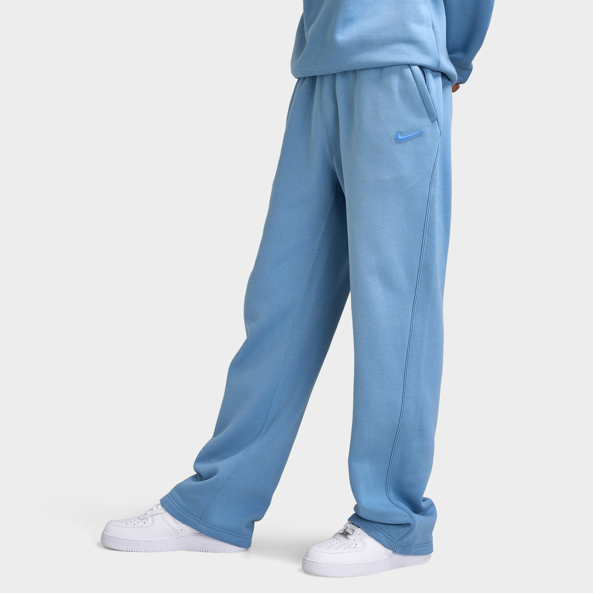 Nike NOCTA Fleece Open Hem Pants. Side view of model wearing work blue sweatpants with relaxed legs and open hems.