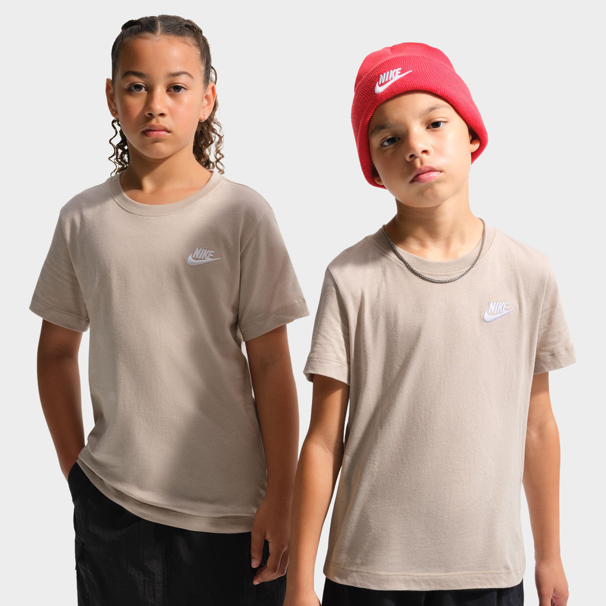Nike Juniors' Futura T-Shirt. Half-length front view of two youth models wearing matching shortsleeve cotton t-shirts in moon particle beige, each featuring a small white embroidered Nike Futura logo on the left chest and a ribbed crewneck collar. 
