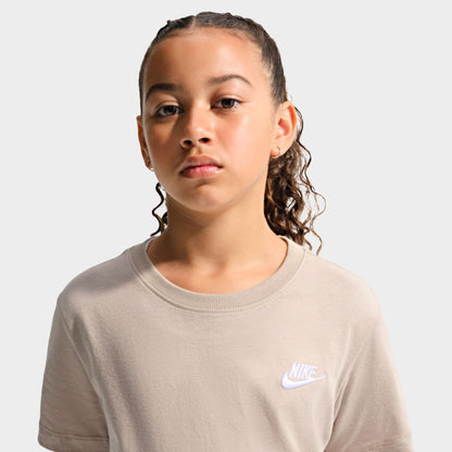 Nike Juniors' Futura T-Shirt. Medium close-up front view of a youth model wearing the moon particle beige t-shirt, displaying the small white embroidered Nike Futura chest logo and ribbed crewneck. 