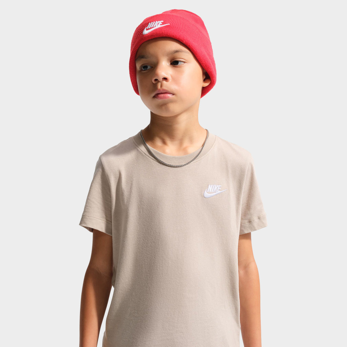 Nike Juniors' Futura T-Shirt. Medium close-up front view of a youth model wearing a red beanie and the moon particle beige t-shirt, displaying the small white embroidered Nike Futura chest logo. 