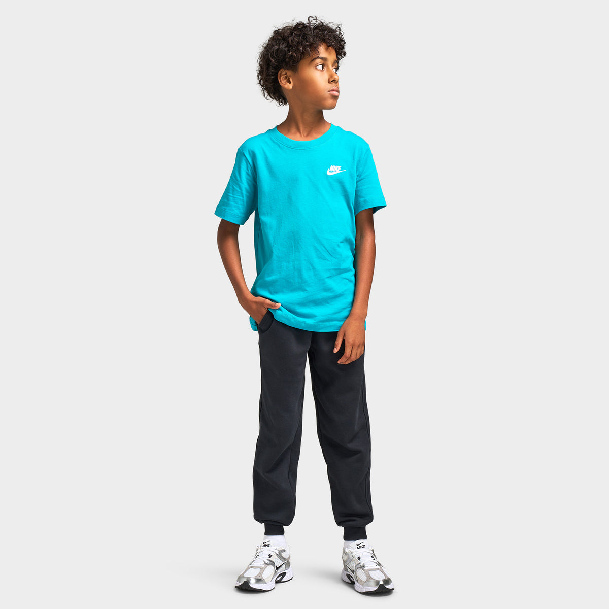 Nike Juniors' Futura T-Shirt. Full-length front model view styled with dark grey joggers and white athletic sneakers. 
