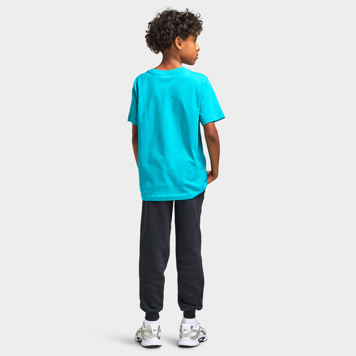 Nike Juniors' Futura T-Shirt. Full-length rear model view showing the back profile of the energy green turquoise shortsleeve t-shirt paired with dark grey joggers and white sneakers. 