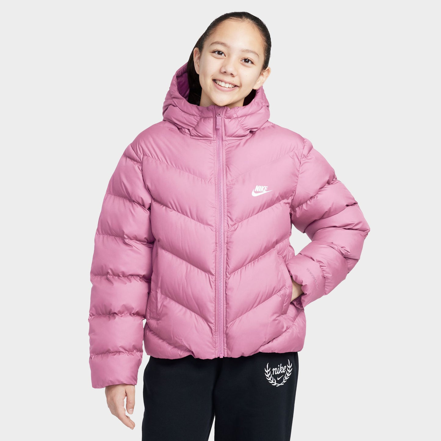 Nike Junior Girls' Therma-FIT Loose-Fit Puffer Jacket. Front view of model wearing pink rise puffer jacket with hood, quilted panels and white Nike Swoosh.