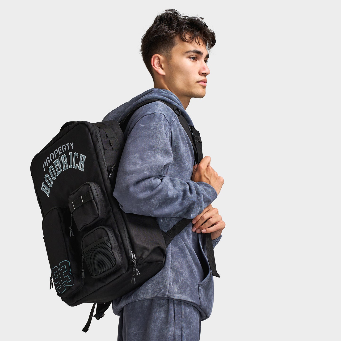 Hoodrich Varso Backpack. Three-quarter view of model wearing black backpack with multiple compartments, padded shoulder straps and large Hoodrich branding.