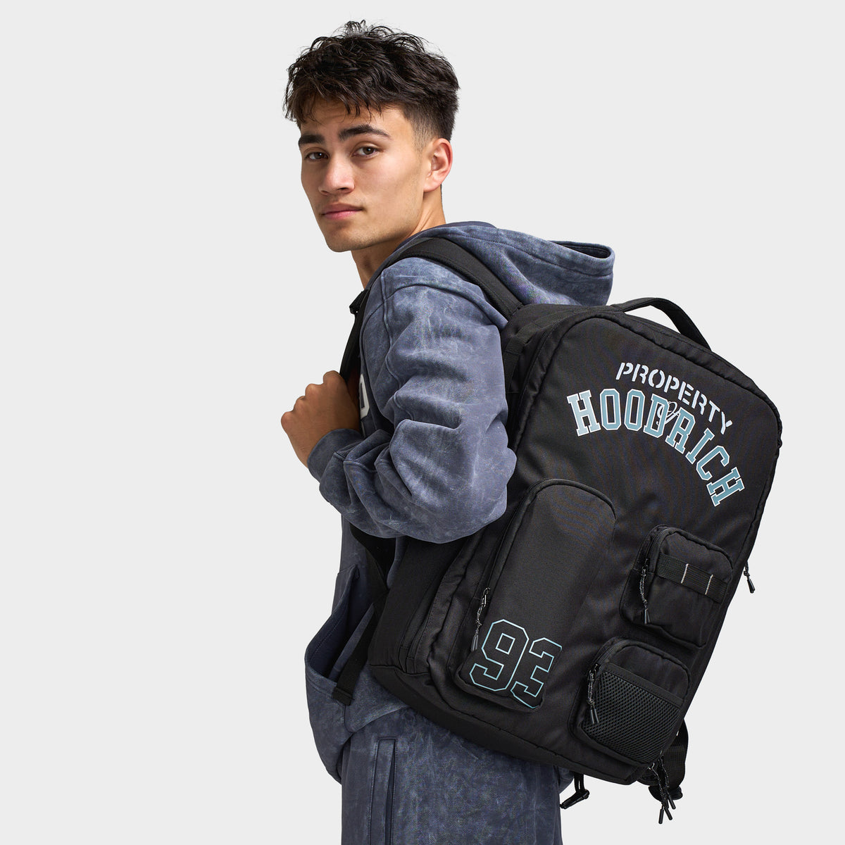 Hoodrich Varso Backpack. Side view of model wearing black backpack with padded shoulder straps, front pockets and Hoodrich branding.