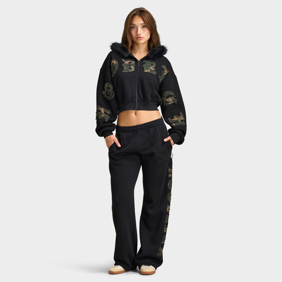 Hoodrich Women's Camo Rift Boxy Zip-Through Hoodie. Full-length front view of the model wearing the black cropped camo zip-through hoodie paired with matching black track pants. 