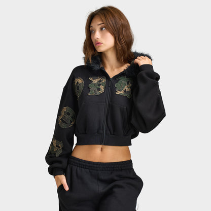 Hoodrich Women's Camo Rift Boxy Zip-Through Hoodie. Front three-quarter view of a female model wearing a black cropped boxy zip-up hoodie featuring woodland camouflage gothic branding appliqué across the chest and right sleeve, faux-fur trimmed hood lining, full-length front zipper, and ribbed cropped hem. 