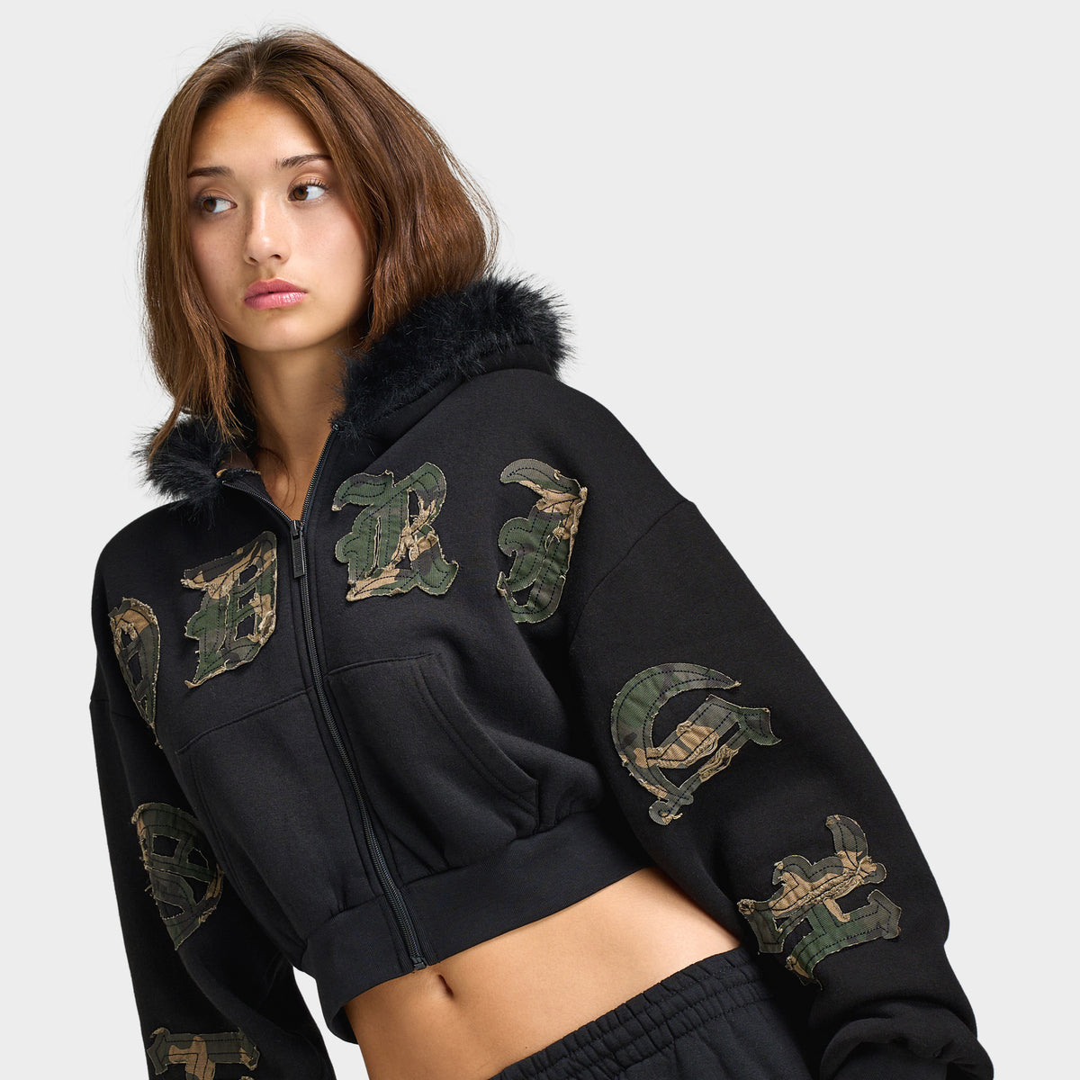 Hoodrich Women's Camo Rift Boxy Zip-Through Hoodie. Angled front view highlighting the black zip hoodie, camouflage logo appliqués across the chest and sleeve, faux-fur hood accent, and boxy cropped fit. 