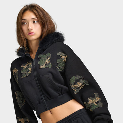 Hoodrich Women's Camo Rift Boxy Zip-Through Hoodie. Angled front view highlighting the black zip hoodie, camouflage logo appliqués across the chest and sleeve, faux-fur hood accent, and boxy cropped fit. 