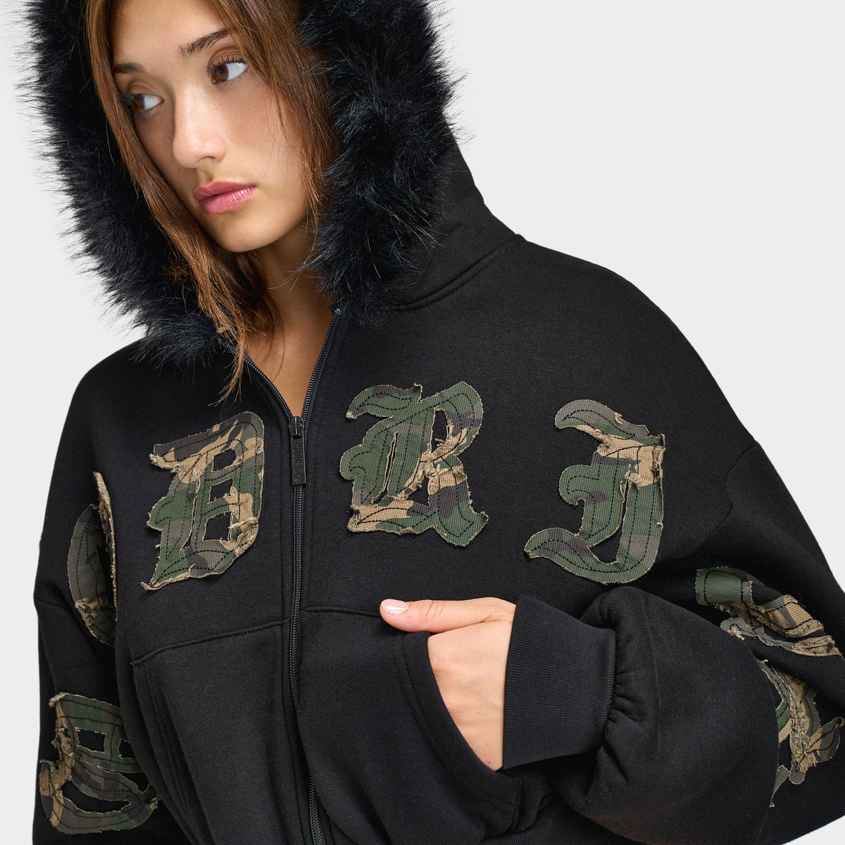 Hoodrich Women's Camo Rift Boxy Zip-Through Hoodie. Close-up view of the model wearing the plush black faux-fur lined hood up, showing the full zip closure, split hand pockets, and camouflage gothic lettering across the chest. 