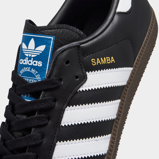 adidas Originals Women's Samba OG. Close-up of core black, white and clear granite sneaker showing black laces, white three stripes, blue tongue label and gold Samba branding.