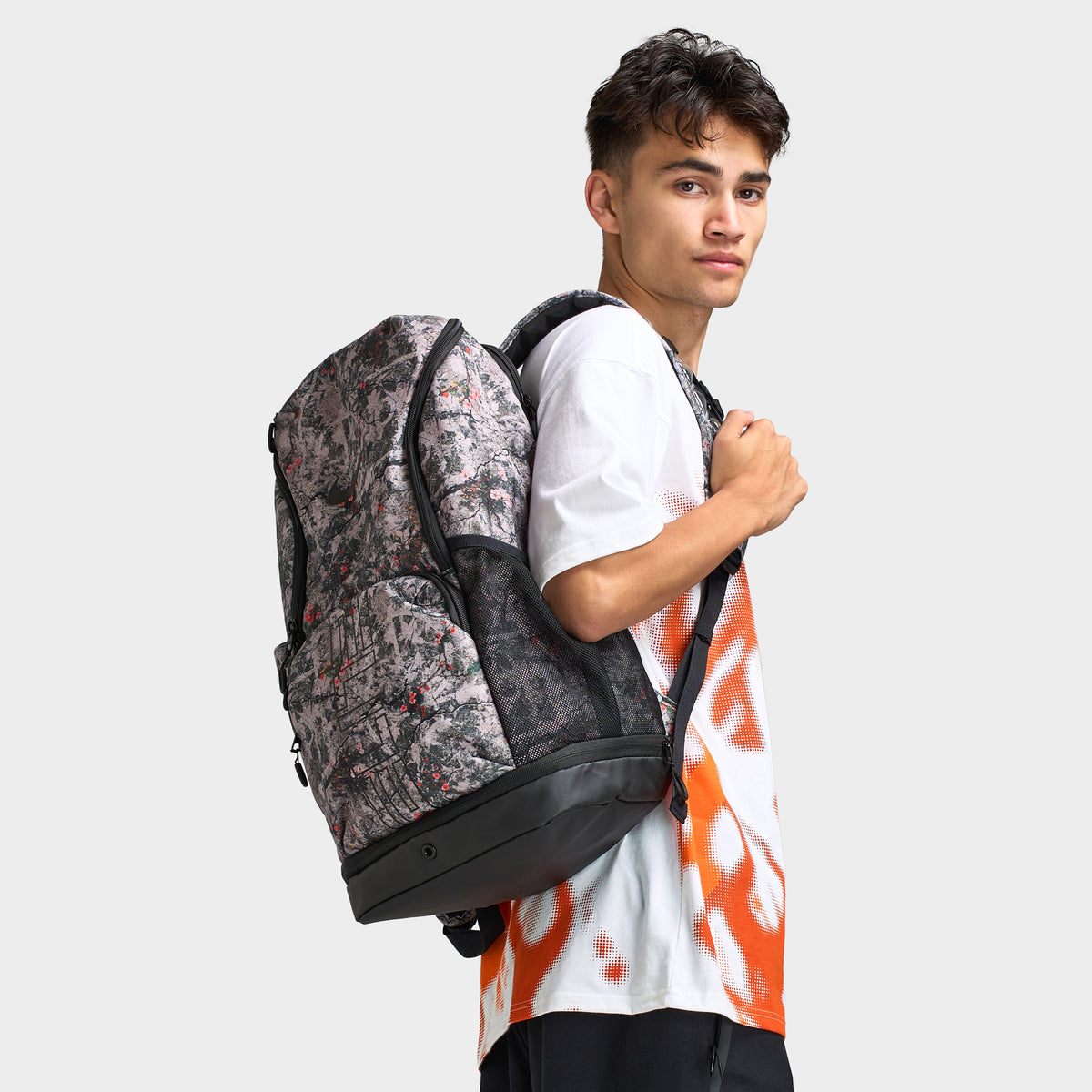 Nike Varsity Elite Backpack. Three-quarter view of model wearing cream and black backpack with all-over graphic print, padded shoulder straps and structured base.