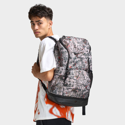 Nike Varsity Elite Backpack. Side view of model wearing cream and black backpack with patterned exterior, multiple compartments and padded straps.