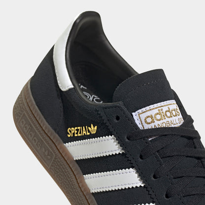 adidas Juniors' Handball Spezial. Close-up of black, white and gum junior sneaker showing black laces, white three stripes, gold Spezial branding and tongue label.