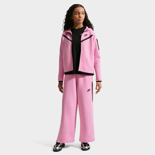 Nike Juniors’ Sportswear Tech Fleece Wide Leg Pant. Full-length front model view of pink rise Tech Fleece wide-leg pants featuring an elastic waistband, black printed Nike Futura logo on the left thigh, vertical zippered side pocket surrounded by black tape, and wide open-leg hems, styled with a matching pink rise full-zip hoodie. 