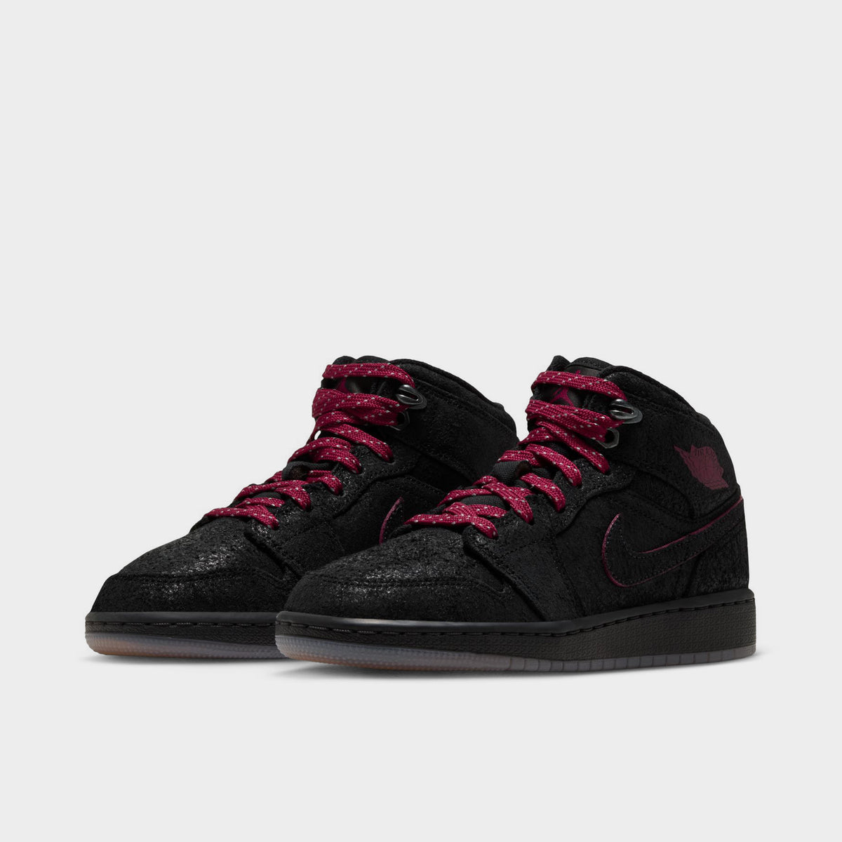 Jordan 1 Mid SE GS. Three-quarter view of pair displaying black textured upper, team red patterned laces, metallic top eyelets, and gum outsoles. 