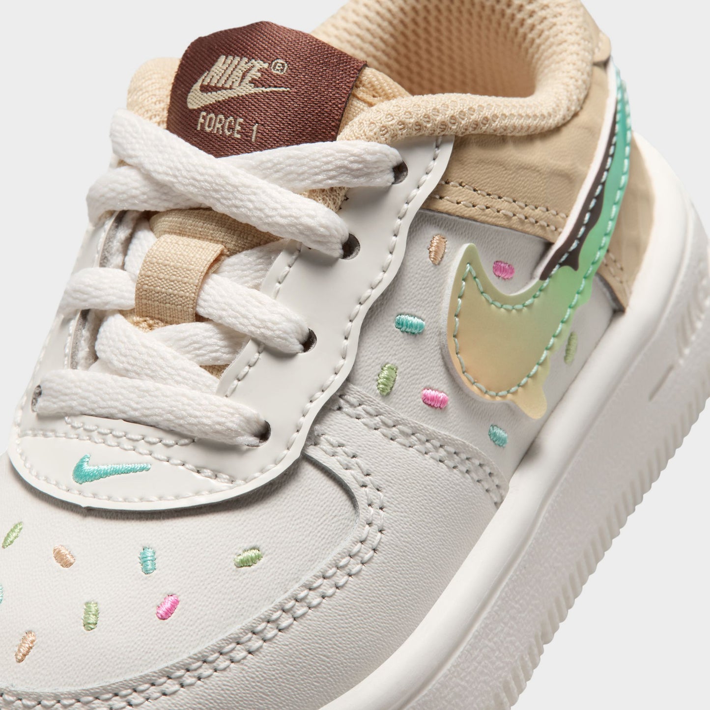 Nike Air Force 1 TD. Close-up of sail, multicolour and sesame toddler sneaker showing white laces, brown tongue label, pastel Swoosh and colourful perforations.