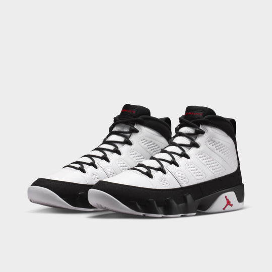 Jordan 9 Retro. Three-quarter view of a pair of high-top basketball shoes showing white leather uppers, black mudguards, black laces, black collars with true red tongue branding, and white outsoles. 