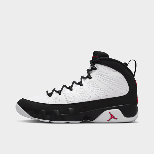 Jordan 9 Retro. Side view of high-top basketball shoes featuring a white leather upper with perforated side panels, black mudguard and midsole overlay, black round laces, black speed lacing eyelets, high-cut black collar lining, black heel pull tab, and a white rubber sole with a true red Jumpman logo at the lateral heel. 