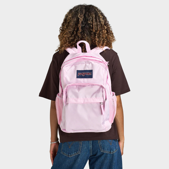 JanSport Big Plans Backpack. Back view of model wearing pink backpack with padded shoulder straps, front zip pocket and JanSport logo patch.