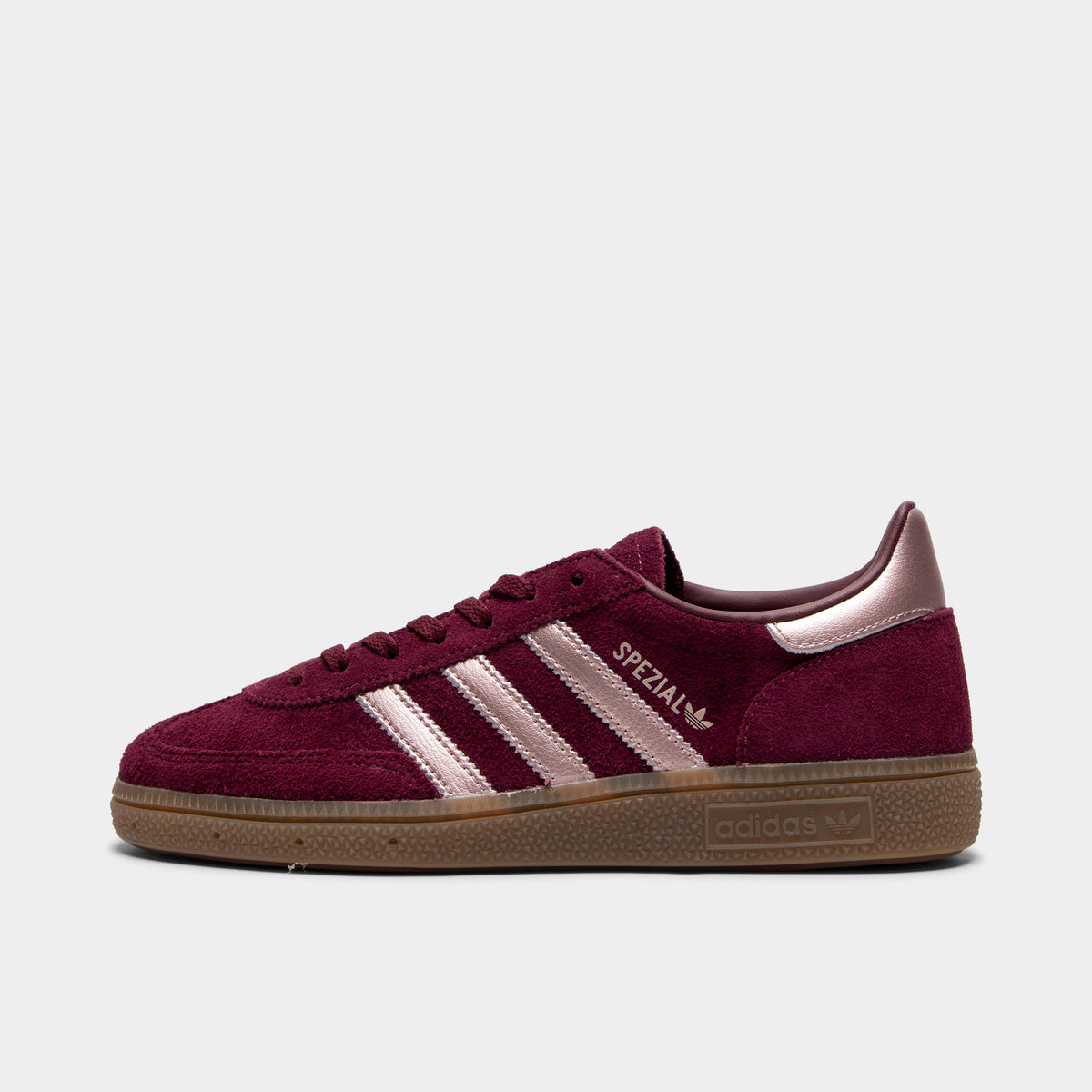 adidas Women's Handball Spezial. Side view of low-top sneakers featuring a maroon suede upper, vapour grey metallic serrated 3-Stripes on the midfoot, vapour grey "SPEZIAL" and Trefoil text branding, metallic vapour grey heel tab, maroon laces, and a gum rubber sole with embossed adidas branding. 