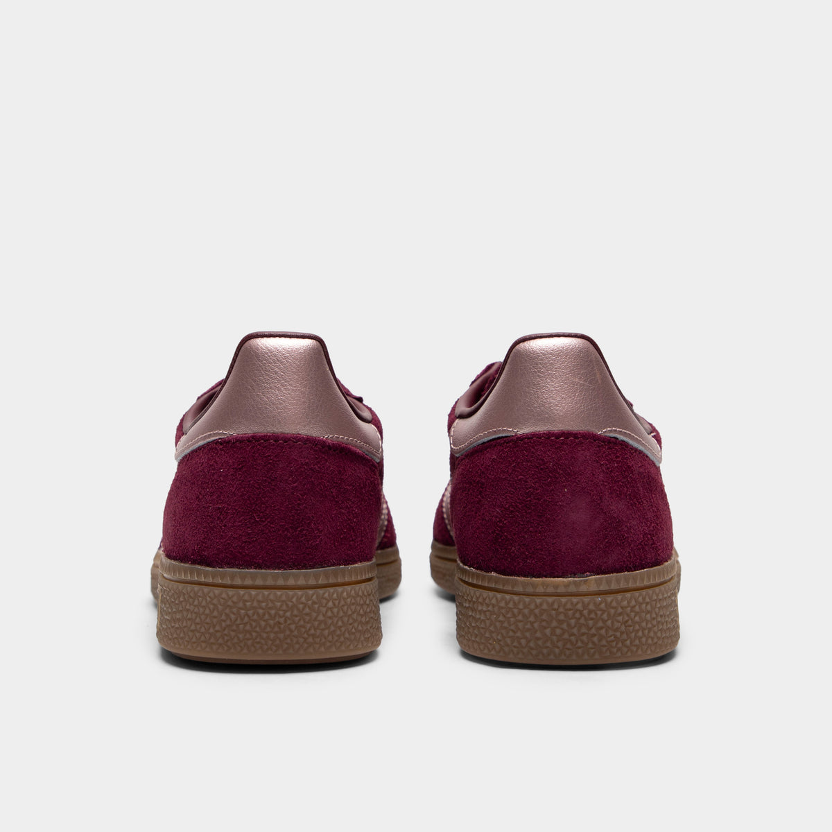 adidas Women's Handball Spezial. Rear view of a pair of low-top sneakers displaying maroon suede heel counters, metallic vapour grey leather heel tabs, and low-profile gum rubber soles. 