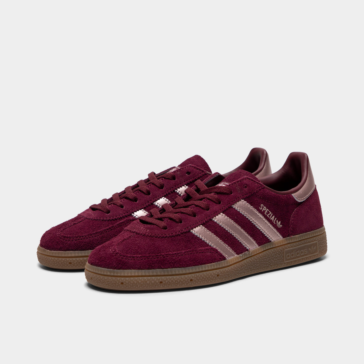 adidas Women's Handball Spezial. Three-quarter view of a pair of low-top sneakers showing maroon suede uppers, vapour grey metallic 3-Stripes, metallic heel tabs, maroon laces, and gum rubber soles. 