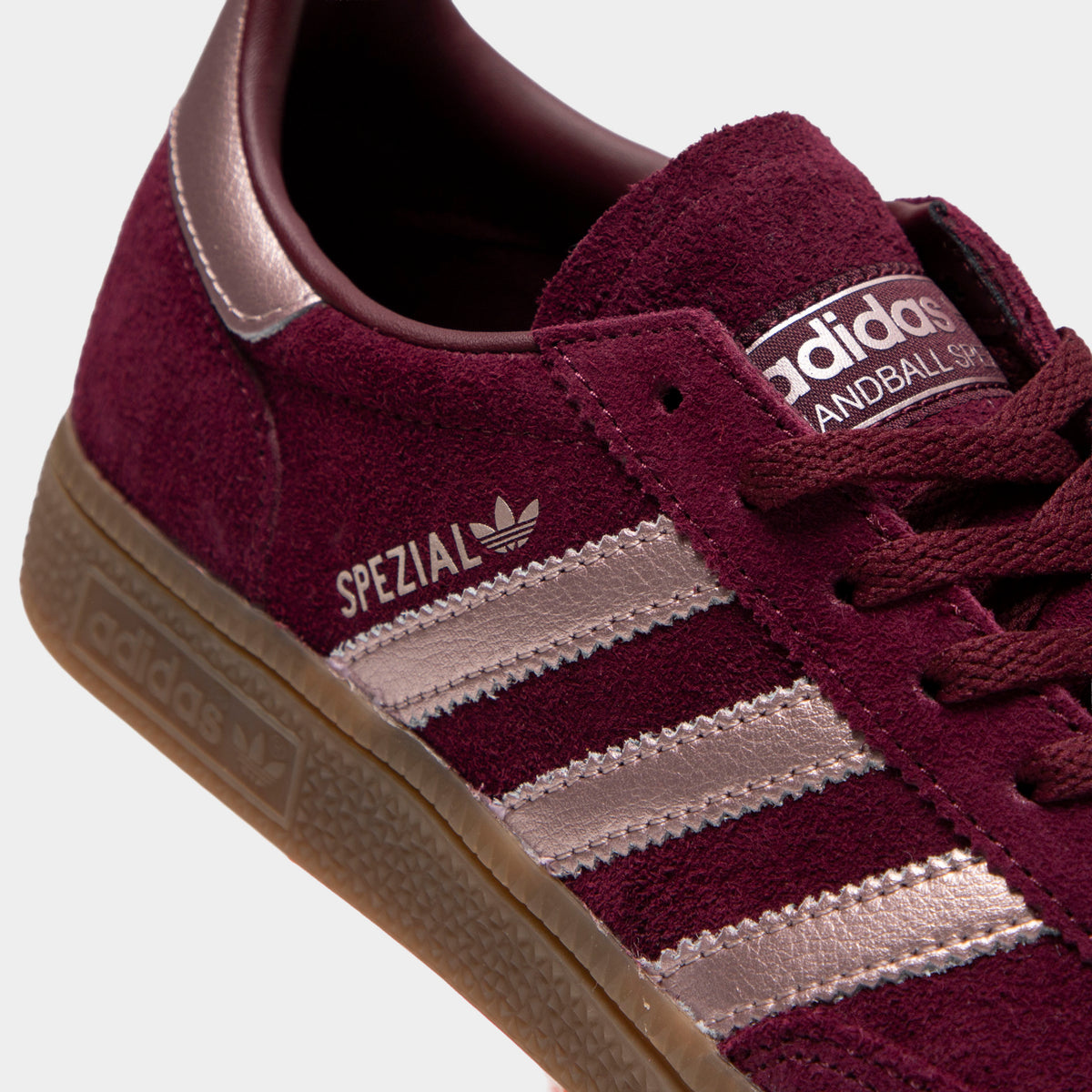 adidas Women's Handball Spezial. Close-up view of tongue and lacing highlighting maroon suede construction, metallic vapour grey serrated 3-Stripes, gold foil "SPEZIAL" text logo, maroon laces, and suede tongue patch with printed adidas branding. 