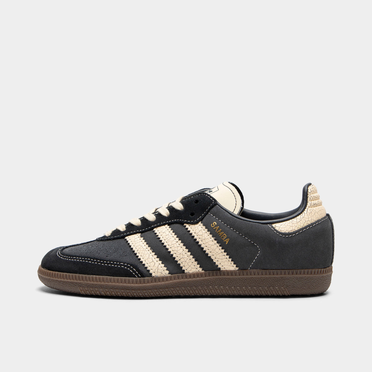 adidas Juniors' Samba OG. Side view of low-top sneakers featuring a carbon dark grey suede upper with black suede T-toe overlay, warm vanilla off-white cracked leather serrated 3-Stripes on the midfoot, gold foil "Samba" text branding, cracked leather warm vanilla heel tab, warm vanilla laces, warm vanilla leather tongue, and a gum rubber sole. 