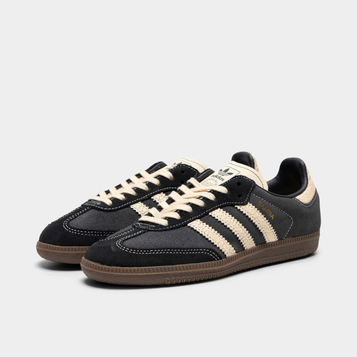 adidas Juniors' Samba OG. Three-quarter view of a pair of low-top sneakers showing carbon dark grey suede uppers, cracked leather warm vanilla serrated 3-Stripes, warm vanilla laces, cracked leather warm vanilla heel tabs, and gum rubber soles. 